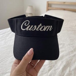 May include: A black visor featuring the word Custom embroidered in white script lettering across the front panel. The visor has a structured brim and an open top design, providing a clear view of the fabric texture and the white stitching detail.