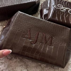 May include: Three dark brown rectangular pouches with a textured crocodile skin pattern. Each pouch features embroidered monogram lettering on the front. One pouch displays the letters AJM in dark brown thread, while two others show white script initials.