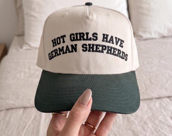 May include: A beige baseball cap with a dark green brim. The front of the cap features the embroidered black text "HOT GIRLS HAVE GERMAN SHEPHERDS" in a bold, arched font. The cap has a structured crown with a small dark button at the top center.