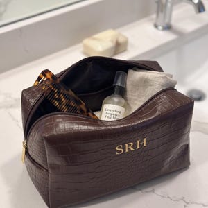 May include: A dark brown, crocodile-textured toiletry bag with a gold zipper and the gold-lettered monogram SRH. The open bag contains a tortoiseshell comb, a clear spray bottle labeled Lavender & Bergamot Face Mist, and a folded beige cloth.