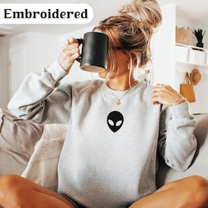 Peut inclure: Un sweat-shirt gris clair avec un motif de tête d'extraterrestre noir. Le mot "Embroidered" est imprimé en blanc. Le sweat-shirt a des manches longues et un col rond.