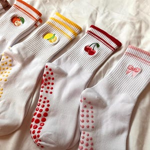 May include: Four pairs of white socks with colorful accents and fruit embroidery. Each pair features a different design: an orange, a lemon, cherries, and a pink bow. The socks have colored stripes at the top and non-slip grips on the soles.