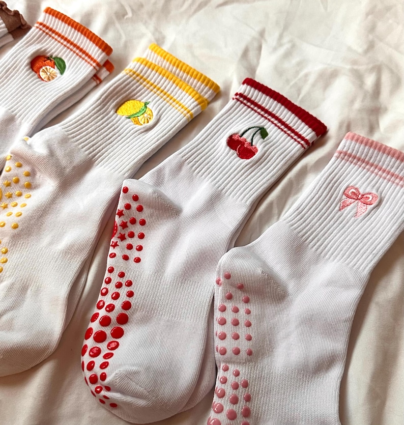 May include: Four pairs of white socks with colorful accents and fruit embroidery. Each pair features a different design: an orange, a lemon, cherries, and a pink bow. The socks have colored stripes at the top and non-slip grips on the soles.
