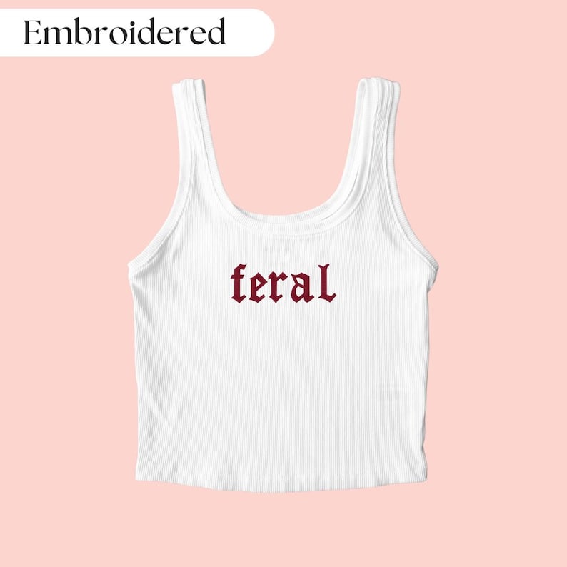May include: White ribbed tank top with the word "feral" embroidered in burgundy gothic lettering. The word "Embroidered" is in a white rounded rectangle in the top left corner. The tank top is on a light pink background.