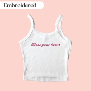 May include: White vest top with thin straps. The phrase "Bless your heart" is embroidered in a cursive font across the front. The word "Embroidered" is above the vest top.