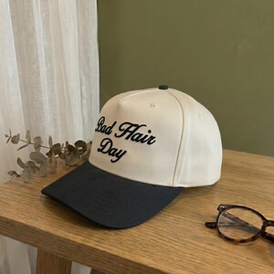 May include: A cream-colored baseball cap with a black brim. The phrase "Bad Hair Day" is embroidered in black script on the front of the cap. The cap is sitting on a wooden surface.