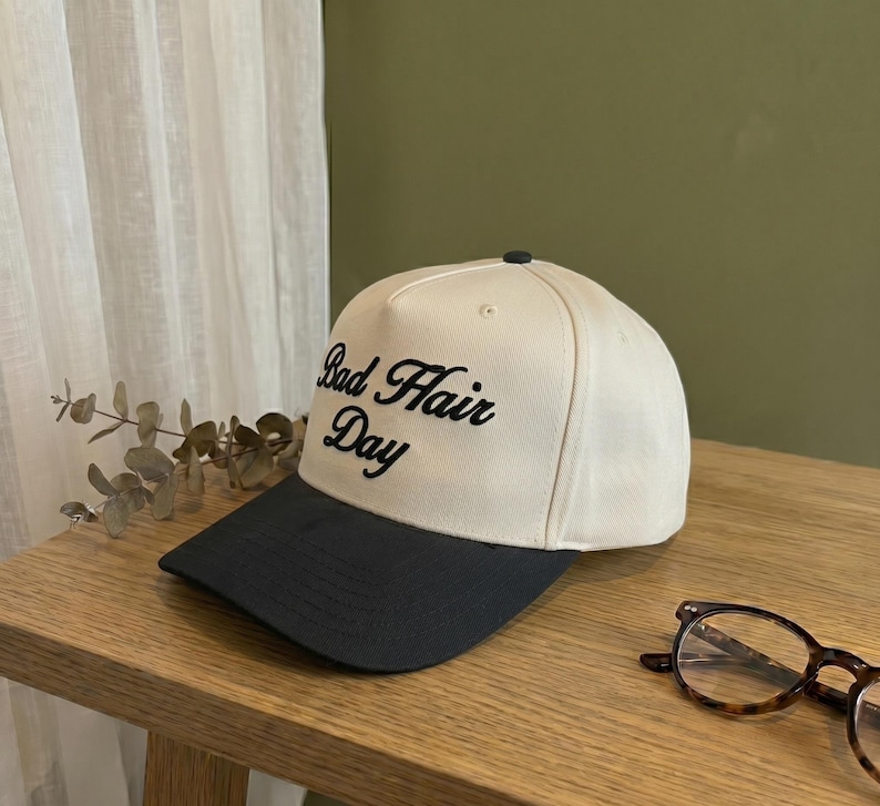 May include: A cream-colored baseball cap with a black brim. The phrase "Bad Hair Day" is embroidered in black script on the front of the cap. The cap is sitting on a wooden surface.