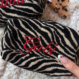 Puede incluir: Un neceser rectangular con estampado de rayas de cebra en negro y beige, con el nombre Gabi bordado en letra roja en la parte frontal. Un segundo neceser a juego y una pinza para el pelo de carey son visibles en el fondo.