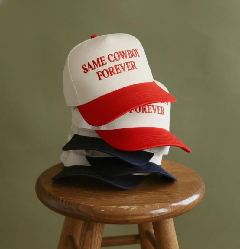 Puede incluir: Una pila de gorras de b&eacute;isbol con el texto "SAME COWBOY FOREVER" en rojo. Las gorras tienen una corona blanca y una visera roja. Una gorra tiene una visera azul marino. Las gorras est&aacute;n apiladas sobre un taburete de madera.