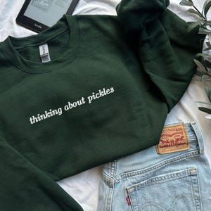 Peut inclure: Un sweat-shirt vert foncé avec le texte "thinking about pickles" brodé en blanc. Le sweat-shirt est plié et posé sur une surface blanche avec un jean bleu.