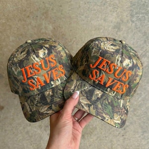 May include: Two camouflage baseball caps with the embroidered text "JESUS SAVES" in orange. The caps have a classic six-panel design and a curved brim. The background is a neutral, light-colored surface.