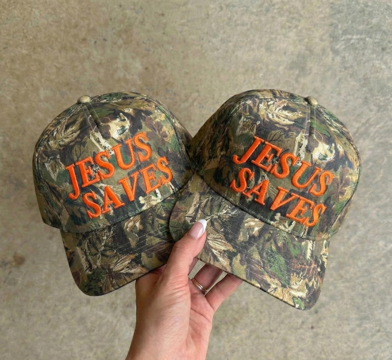 May include: Two camouflage baseball caps with the embroidered text "JESUS SAVES" in orange. The caps have a classic six-panel design and a curved brim. The background is a neutral, light-colored surface.
