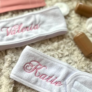 Op de afbeelding: Twee witte badstof haarbanden met roze geborduurde namen, "Valeria" en "Katie". De haarbanden liggen op een zacht, crèmekleurig oppervlak, met andere schoonheidsproducten op de achtergrond.