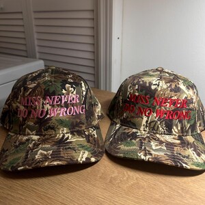 May include: Two camouflage baseball caps with the embroidered text "MISS NEVER DO NO WRONG" in pink and red. The caps feature a classic six-panel design with a curved brim, suitable for outdoor activities or casual wear.