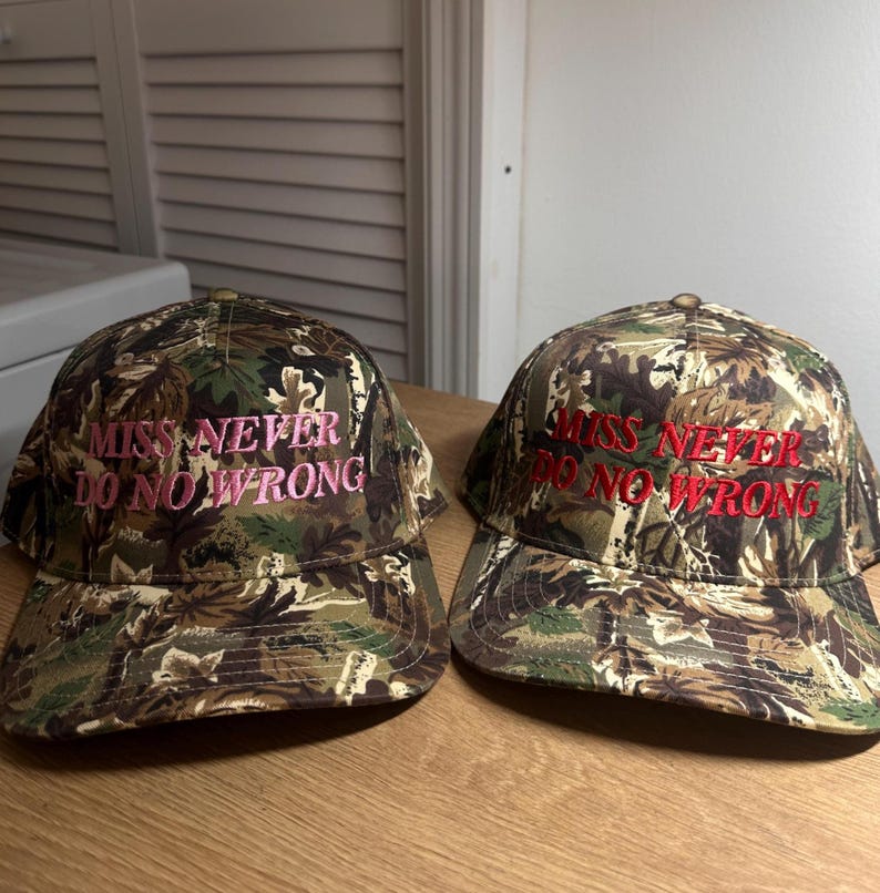 May include: Two camouflage baseball caps with the embroidered text "MISS NEVER DO NO WRONG" in pink and red. The caps feature a classic six-panel design with a curved brim, suitable for outdoor activities or casual wear.