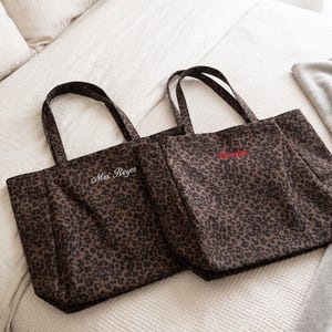 Op de afbeelding: Twee rechthoekige tote bags met een bruin en zwart luipaardpatroon. De linker tas heeft de tekst Mrs. Reyes in het wit geborduurd, terwijl de rechter tas rode geborduurde tekst bevat. Beide tassen hebben twee bijpassende stoffen schouderbanden.
