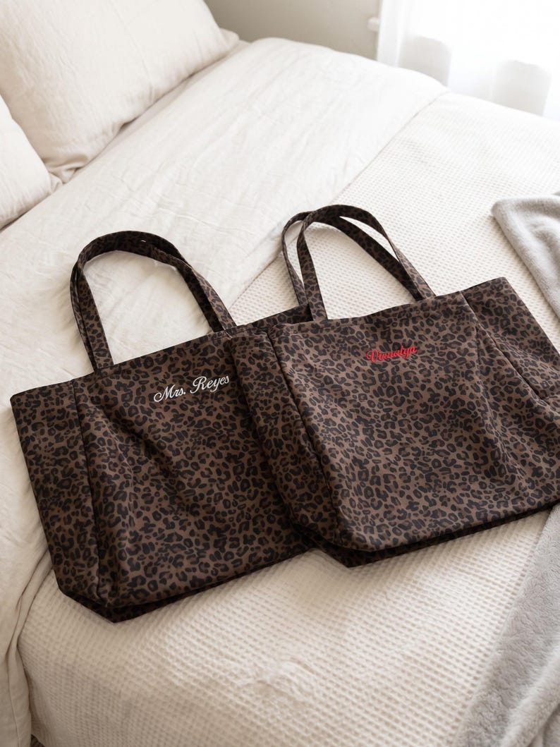 Op de afbeelding: Twee rechthoekige tote bags met een bruin en zwart luipaardpatroon. De linker tas heeft de tekst Mrs. Reyes in het wit geborduurd, terwijl de rechter tas rode geborduurde tekst bevat. Beide tassen hebben twee bijpassende stoffen schouderbanden.