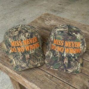 May include: Two camouflage baseball caps with the text "MISS NEVER DO NO WRONG" embroidered in orange. The caps are made of a fabric with a pattern of green, brown, and tan. They are sitting on a weathered wooden surface.
