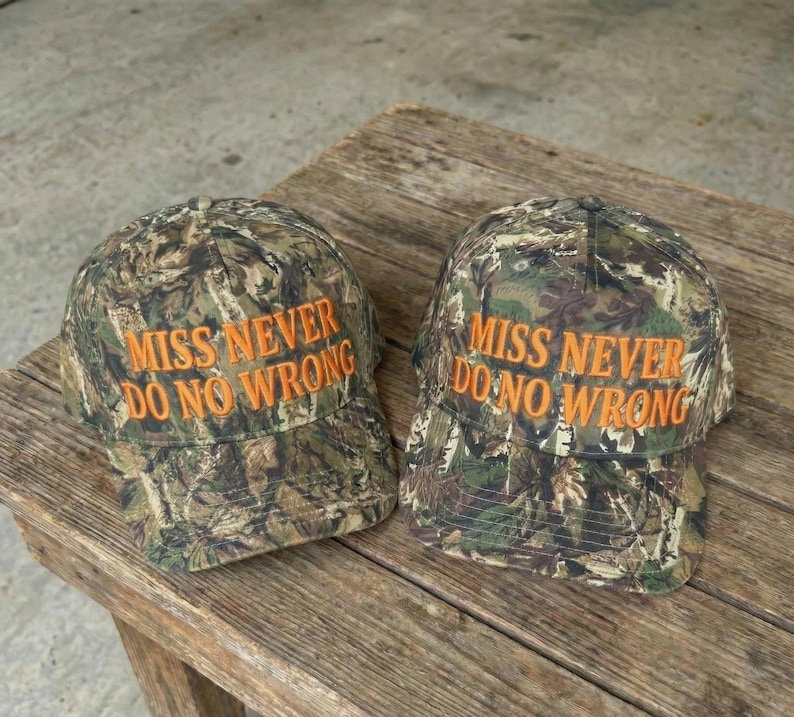 May include: Two camouflage baseball caps with the text "MISS NEVER DO NO WRONG" embroidered in orange. The caps are made of a fabric with a pattern of green, brown, and tan. They are sitting on a weathered wooden surface.