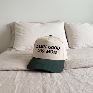 May include: A baseball cap with a cream-coloured crown and a dark green brim. The text "DAMN GOOD DOG MOM" is printed in black across the front. The cap rests on a bed with cream-coloured bedding.