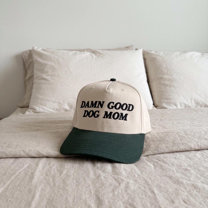 May include: A baseball cap with a cream-coloured crown and a dark green brim. The text "DAMN GOOD DOG MOM" is printed in black across the front. The cap rests on a bed with cream-coloured bedding.