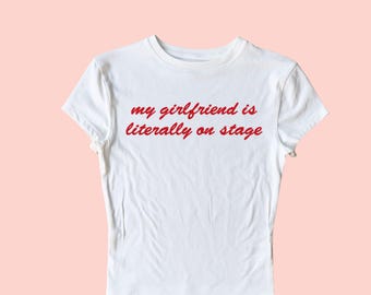 Könnte beinhalten: Weißes T-Shirt mit rotem Sticktext, der "my girlfriend is literally on stage" lautet.