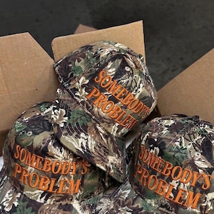 Pode incluir: Bonés de beisebol camuflados com o texto bordado "SOMEBODY'S PROBLEM" em laranja. Os bonés estão numa caixa de cartão, sugerindo que estão prontos para venda ou envio. Os bonés têm um design clássico de seis painéis.