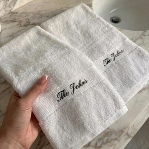 May include: Two white, fluffy hand towels with the embroidered text "The Johns" in black script. The towels are folded and placed on a marble countertop near a white sink. The towels are made of a soft, absorbent material.