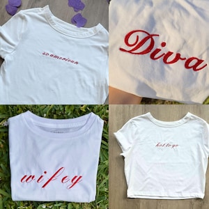 Op de afbeelding: Vier witte t-shirts met rood geborduurde tekst. De shirts tonen de woorden "so american", "Diva", "wifey" en "hot to go". De shirts worden gepresenteerd op een wit oppervlak en op groen gras.