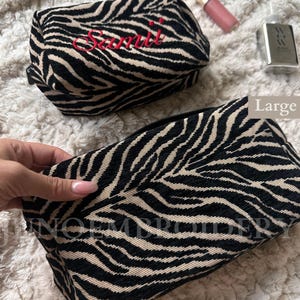 Puede incluir: Dos neceseres rectangulares con un patr&oacute;n de rayas de cebra en negro y beige. El bolso m&aacute;s peque&ntilde;o est&aacute; etiquetado como Medium y muestra el nombre bordado en rojo Samii. El bolso m&aacute;s grande est&aacute; etiquetado como Large. Ambos tienen cremallera.