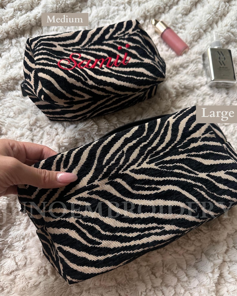 Puede incluir: Dos neceseres rectangulares con un patr&oacute;n de rayas de cebra en negro y beige. El bolso m&aacute;s peque&ntilde;o est&aacute; etiquetado como Medium y muestra el nombre bordado en rojo Samii. El bolso m&aacute;s grande est&aacute; etiquetado como Large. Ambos tienen cremallera.