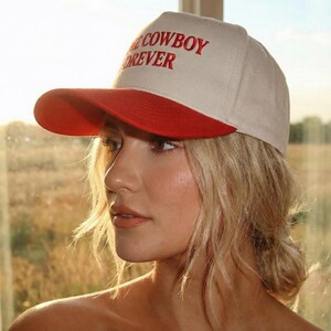 Puede incluir: Gorra de b&eacute;isbol beige con visera roja. El texto "COWBOY FOREVER" est&aacute; bordado en rojo en la parte delantera. La gorra la lleva una persona con cabello rubio.
