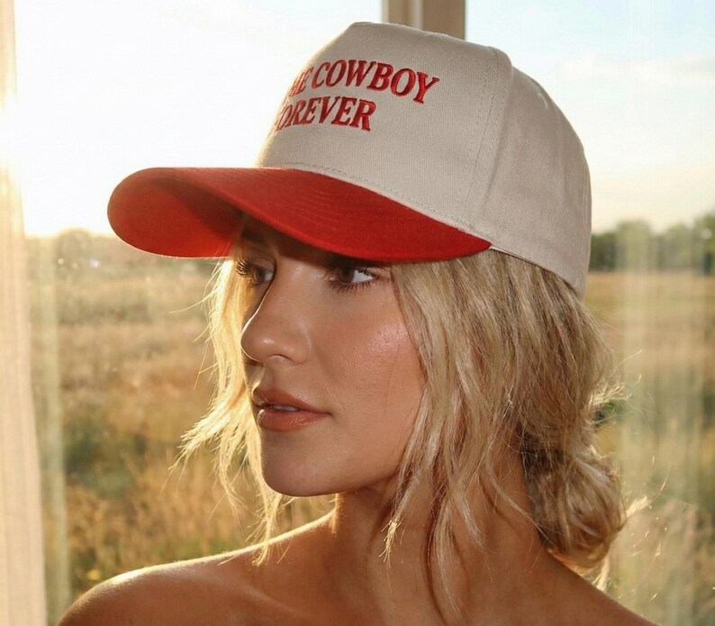 Puede incluir: Gorra de b&eacute;isbol beige con visera roja. El texto "COWBOY FOREVER" est&aacute; bordado en rojo en la parte delantera. La gorra la lleva una persona con cabello rubio.
