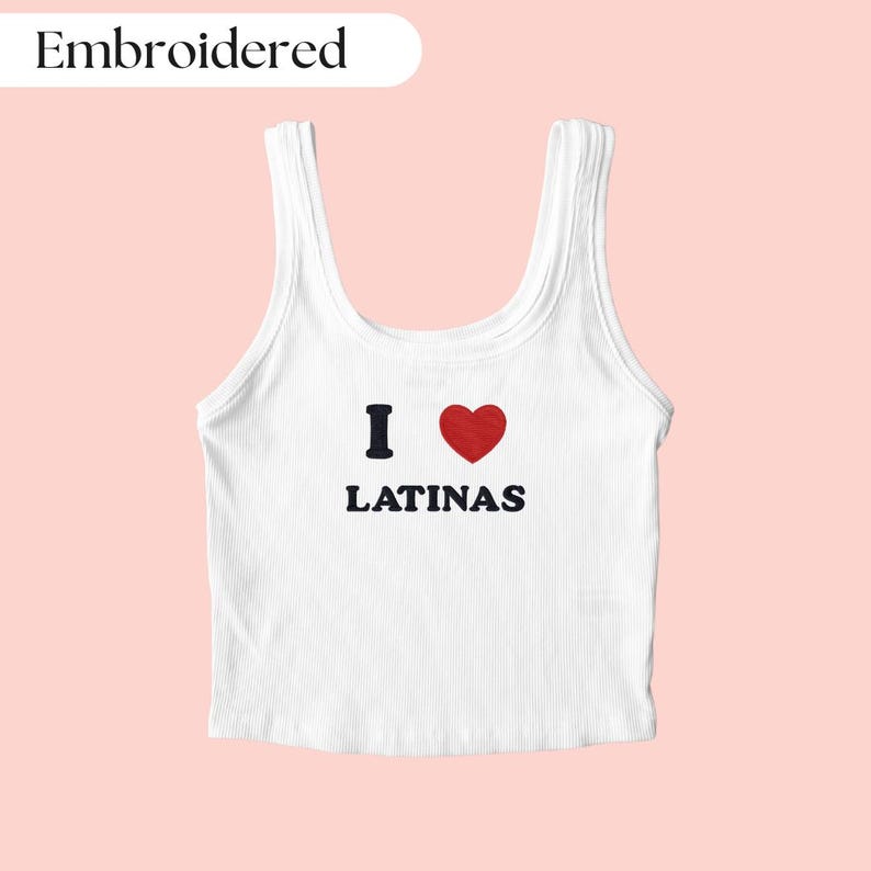 May include: White ribbed vest top with the text "I ❤️ LATINAS" in black and red. The word "Embroidered" is in a white oval in the upper left corner. The background is a solid light pink.