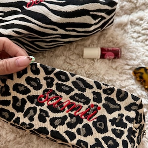 May include: Two cosmetic bags with the name "samii" embroidered in red. One bag has a black and white zebra print, and the other has a tan and black leopard print. A tube of lip gloss is also visible.