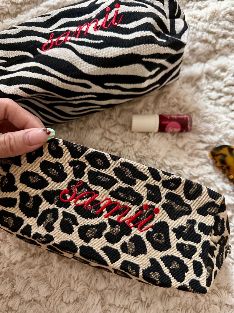 May include: Two cosmetic bags with the name "samii" embroidered in red. One bag has a black and white zebra print, and the other has a tan and black leopard print. A tube of lip gloss is also visible.