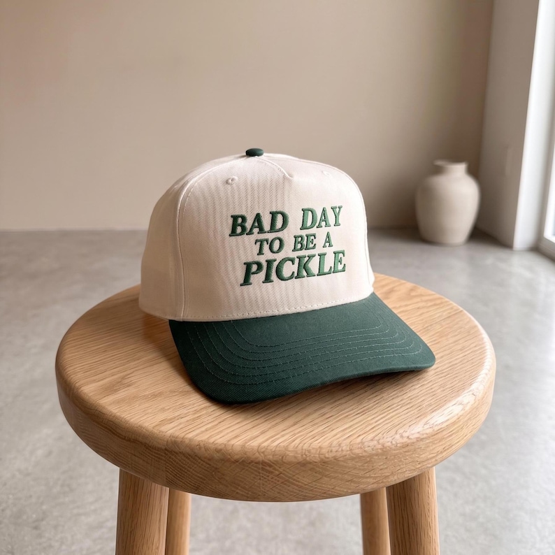 May include: A cream and dark green baseball cap featuring the embroidered phrase "BAD DAY TO BE A PICKLE". The cap has a curved peak and a button detail. It rests on a small wooden stool.
