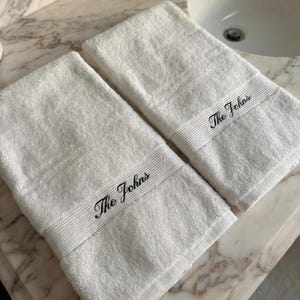 May include: Two white hand towels with the embroidered text "The Johns" in black. The towels are made of a soft, absorbent material and are folded neatly. The towels are placed on a marble surface.