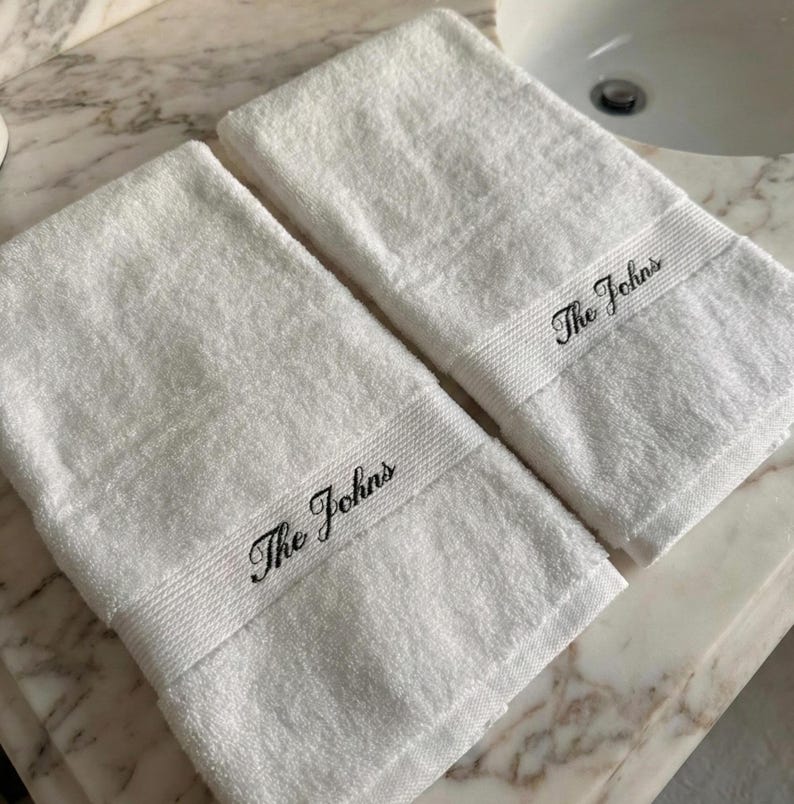 May include: Two white hand towels with the embroidered text "The Johns" in black. The towels are made of a soft, absorbent material and are folded neatly. The towels are placed on a marble surface.