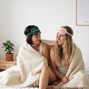 Puede incluir: Dos mujeres con antifaces para dormir y envueltas en una manta beige. Una m&aacute;scara es verde con el texto "sleeping in", la otra es rosa con el texto "dream big". La escena est&aacute; ambientada en un dormitorio con una planta y un cuadro enmarcado.