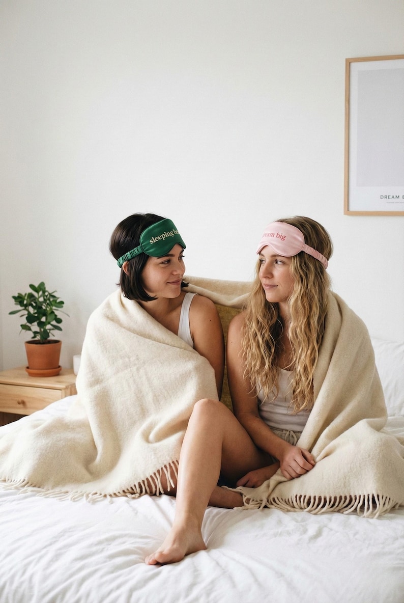 Puede incluir: Dos mujeres con antifaces para dormir y envueltas en una manta beige. Una m&aacute;scara es verde con el texto "sleeping in", la otra es rosa con el texto "dream big". La escena est&aacute; ambientada en un dormitorio con una planta y un cuadro enmarcado.