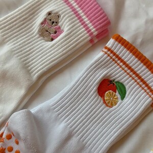 May include: White ribbed socks with embroidered designs. One sock features a teddy bear with a pink scarf, and the other has an orange and lemon design. The sock cuffs have pink and orange stripes.