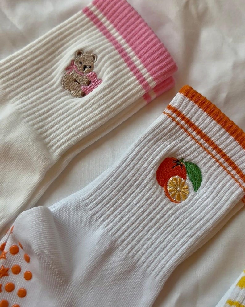 May include: White ribbed socks with embroidered designs. One sock features a teddy bear with a pink scarf, and the other has an orange and lemon design. The sock cuffs have pink and orange stripes.