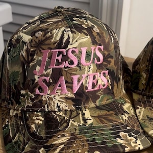 May include: Camouflage baseball cap with the text "JESUS SAVES" embroidered in pink. The cap features a classic six-panel design with a curved brim and adjustable strap. The fabric has a pattern of green, brown, and tan.
