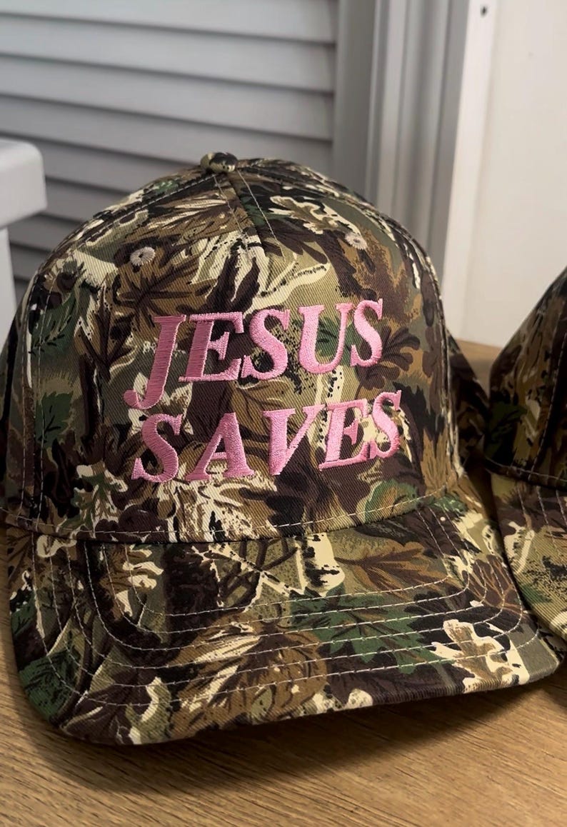 May include: Camouflage baseball cap with the text "JESUS SAVES" embroidered in pink. The cap features a classic six-panel design with a curved brim and adjustable strap. The fabric has a pattern of green, brown, and tan.