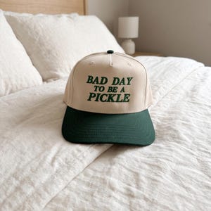 May include: A beige and dark green baseball cap with the text "BAD DAY TO BE A PICKLE" in green. The cap is on a white bedspread. The cap has a dark green peak and a black button on top.