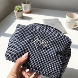 May include: A navy blue and white checkered cosmetic bag with a black zipper. The bag has embroidered initials "P.H." in white script. The bag is held in a person's hand.