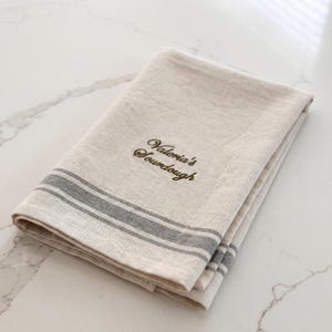 May include: A folded beige linen kitchen towel featuring two horizontal grey stripes near the edge and the embroidered text "Valeria's Sourdough" in a dark brown cursive font centered on the fabric surface.