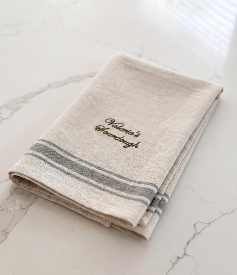 May include: A folded beige linen kitchen towel featuring two horizontal grey stripes near the edge and the embroidered text "Valeria's Sourdough" in a dark brown cursive font centered on the fabric surface.