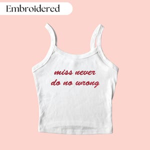 Peut inclure: Débardeur blanc avec fines bretelles, orné de la phrase brodée en rouge "miss never do no wrong". Le mot "Embroidered" est dans un ovale blanc en haut à gauche. Fond rose clair.